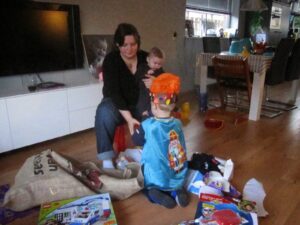 Baby's eerste sinterklaas cadeaus 2 Vrouw met baby op schoot en peuter in Sinterklaas-cape tussen uitgepakte cadeautjes in woonkamer.