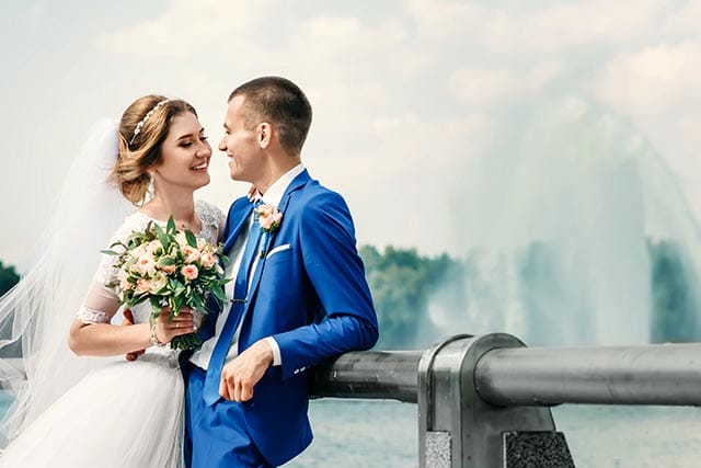 Bruid en bruidegom bij reling met fontein op achtergrond, bruid met boeket, bruidegom in blauw pak.