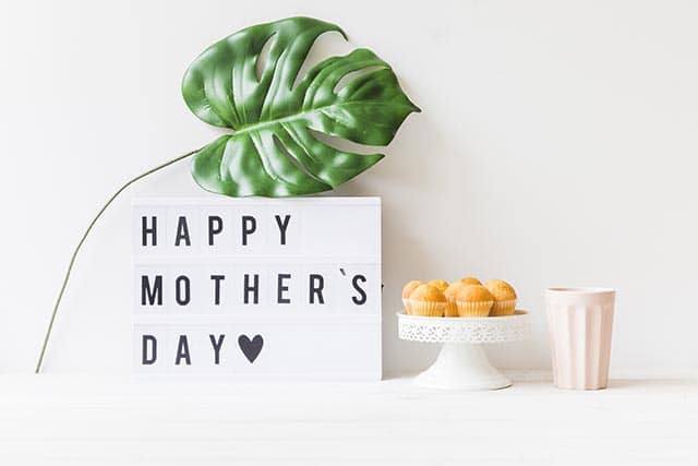 Bord met tekst Happy Mother's Day, groot groen blad, schaal met muffins en lichtroze beker.