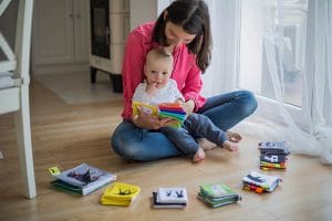 Leesplezier gegarandeerd: de 5 mooiste voorleesboeken! 5 Vrouw zit met baby op houten vloer bij raam, samen kijkend naar kleurrijk stoffen boekje.