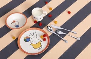 7x de leukste nijntje kraamcadeaus 5 Kinderservies met konijnillustraties, mok met melk, bestek en kleine tomaatjes op gestreept tafelkleed.