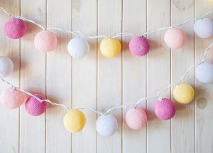 Gekleurde lichtslingers met roze, geel en witte bolletjes tegen een lichte houten achtergrond.