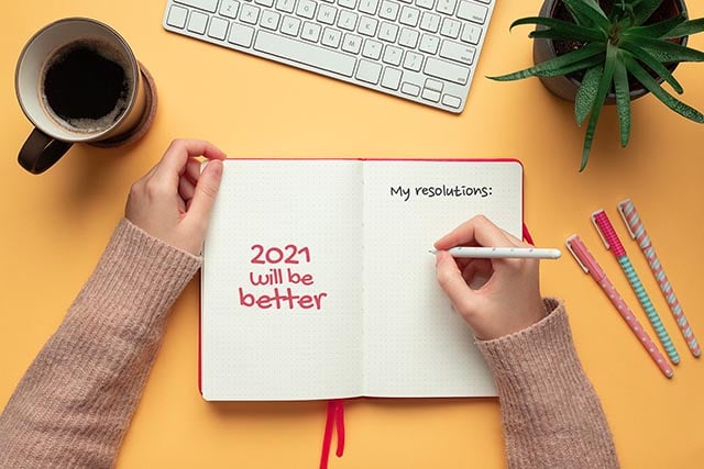 Geef een steuntje in de rug... 2 Hand schrijft in notitieboekje met tekst 2021 will be better op gele ondergrond met koffie, toetsenbord en pennen.