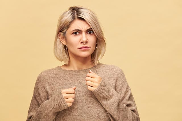 Cadeaus die je niet aan je vriendin moet geven! 2 Vrouw met blond haar en bruine trui kijkt bezorgd met handen omhoog tegen beige achtergrond