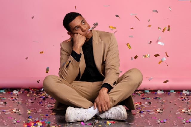 Man in beige pak zit gekruist met feestfluitje en confetti tegen een roze achtergrond.