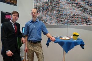 Twee mannen staan bij een tafel met blauwe doek en kleurrijk kunstwerk aan de muur.