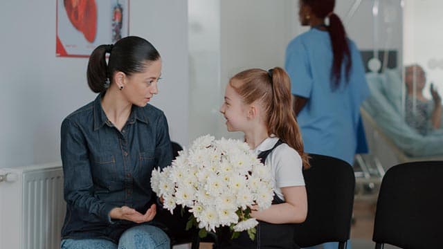 Vrouw en meisje praten terwijl het meisje witte bloemen vasthoudt, verpleegkundige loopt voorbij.