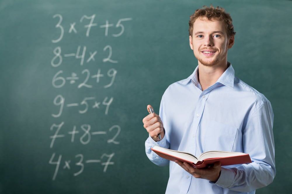 Man met open boek staat voor schoolbord met wiskundesommen en houdt een pen vast.