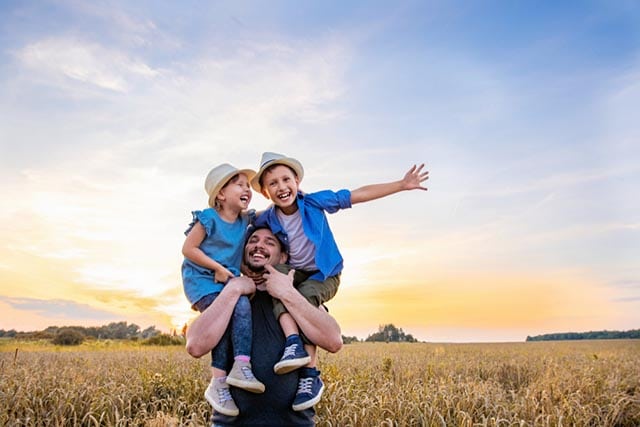 Vader met twee lachende kinderen op zijn schouders in een veld bij zonsondergang