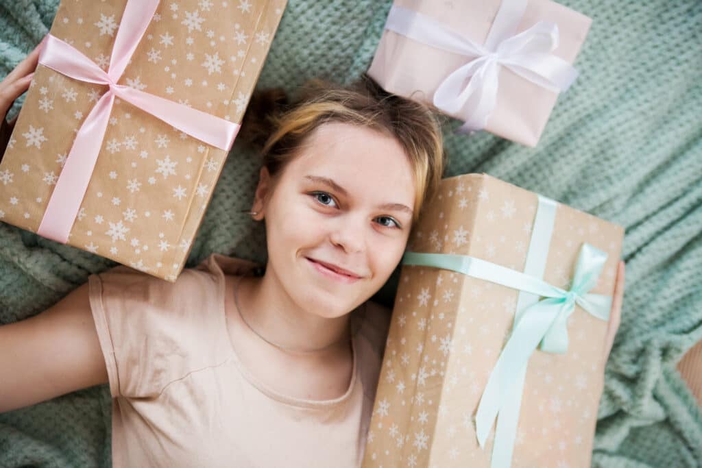 Meisje van 14 jaar ligt omringd door drie ingepakte cadeaus met roze en blauwe strikken op een groen dekbed en kijkt glimlachend omhoog naar de camera