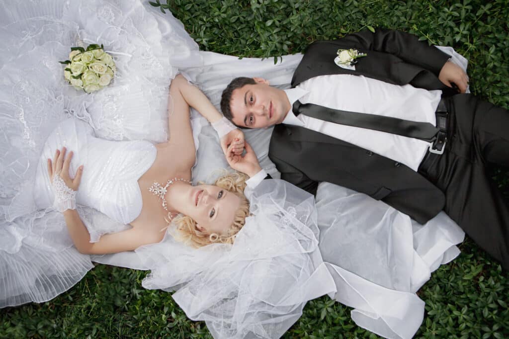 Cadeau voor een bruiloft, wat geef je het bruidspaar cadeau? 2 Bruidspaar ligt hand in hand op groene bladeren, bruid in witte jurk met sluier en bruidegom in zwart pak.