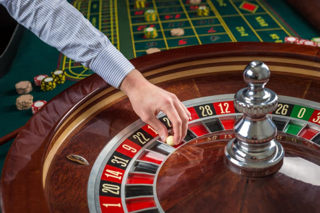 Hand plaatst witte bal op nummer 22 van een roulettewiel in casino met fiches op achtergrond.