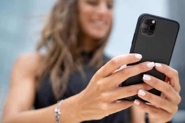 Vrouw met lang haar houdt zwarte smartphone vast en kijkt ernaar met glimlach.