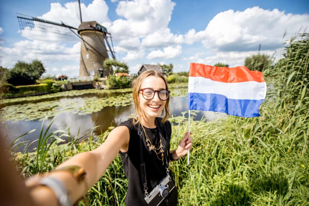Vrouw met camera maakt selfie met Nederlandse vlag bij waterlelies en molen onder blauwe lucht.