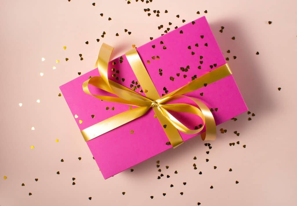 Roze geschenkdoos met gouden lint en hartvormige confetti op lichte achtergrond.